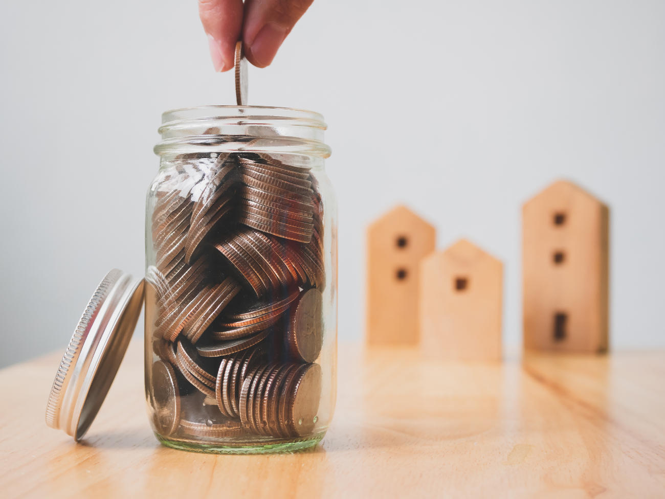 A glass jar filling with money as three wooden toy houses sit in the background.
