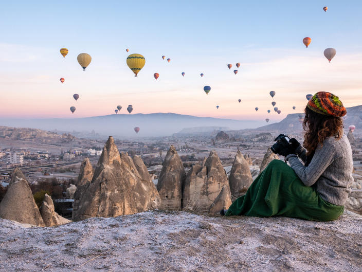 Lady taking pictures of hot air ballons rising over a canyon.