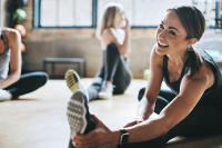 You can become a trainer (or hire a trainer) at All About The Knowledge. Three women stretching in a gym. The most prominent lady is smiling while doing a sitting single leg hamstring stretch.